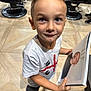 child, boy, book, portrait, floor, tile_floor, barbershop_chair, haircut, white_tshirt, black_shoes, indoor, curious_expression, short_hair, looking_up, standing, hand, person, photography, casual_clothing, real_photo
