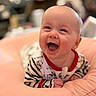baby, infant, child, smiling, laughing, portrait, close_up, indoor, cushion, soft_furnishing, striped_clothing, onesie, red_trim, seated, happy, joyful, face, hands, blurred_background, photography