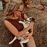 woman, puppy, dog, grass, outdoor, sitting, smiling, glasses, blonde_hair, tattoo, earrings, brown_dress, leash, wooden_bench, happy, playful, pet, nature, sunlight, affection