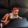 baby, child, toddler, sitting, couch, portrait, smiling, tongue_out, headband, red_clothing, white_shirt, sneakers, black_couch, indoors, furniture, fashion, cute, casual, infant, photography