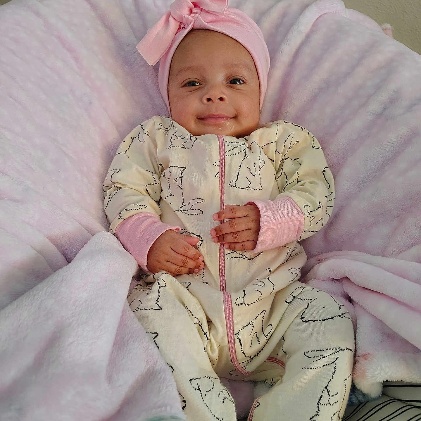 Zoei Zuliana is registered to the contest to win money with this photo: baby, blanket, bow, cheeks, cozy, eyes_open, hands, headband, infant, lying_down, onesie, patterned_clothing, pink, plush, portrait, smiling, soft, tiny_person, wall, zipper