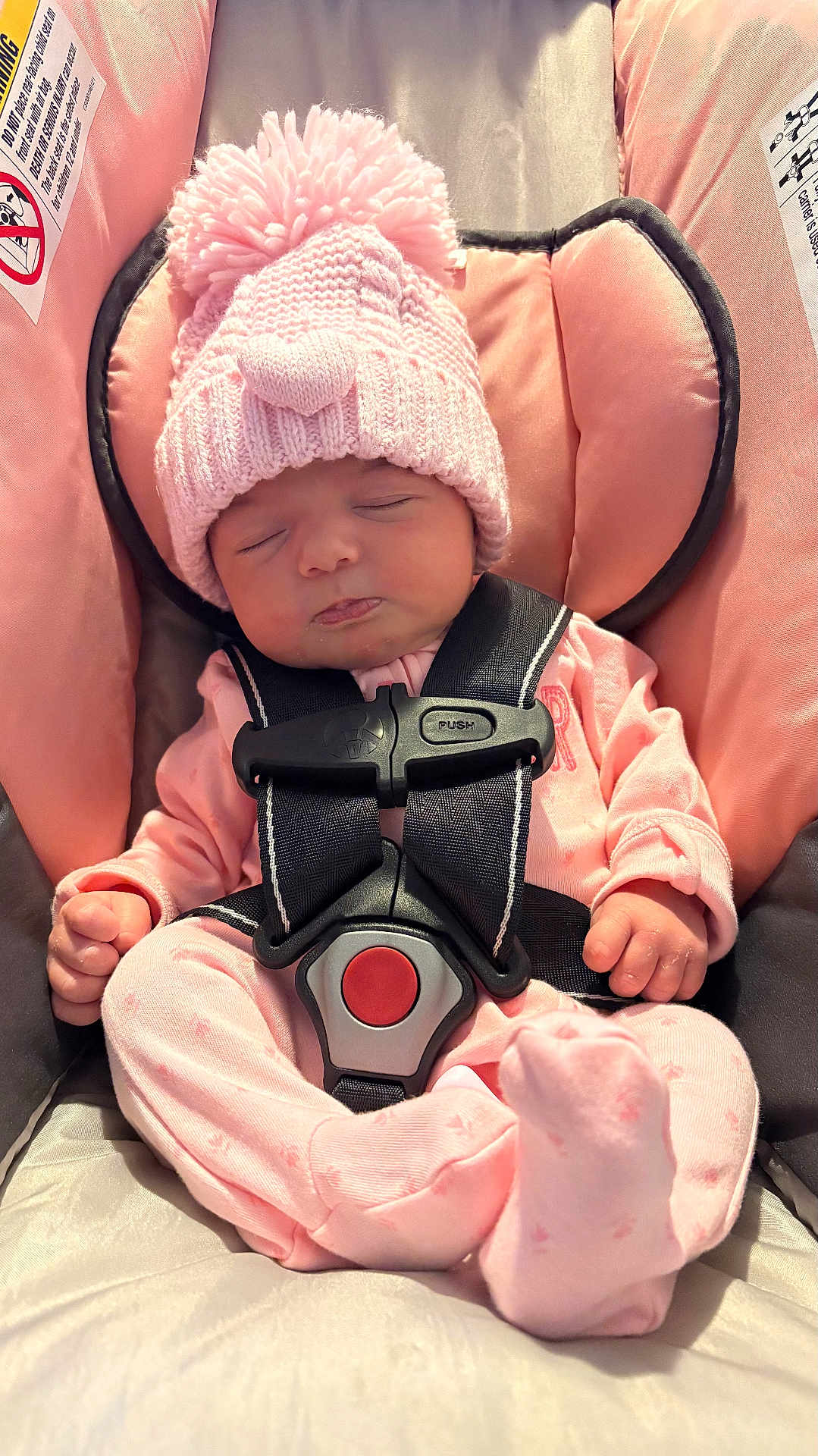 Delilah is registered to the contest to win money with this photo: baby, infant, sleeping, pink_hat, pom_pom, car_seat, safety_harness, pink_outfit, cozy, cute, child, newborn, soft_lighting, indoor, peaceful, closed_eyes, small_hands, footed_pajamas, knit_hat, seat