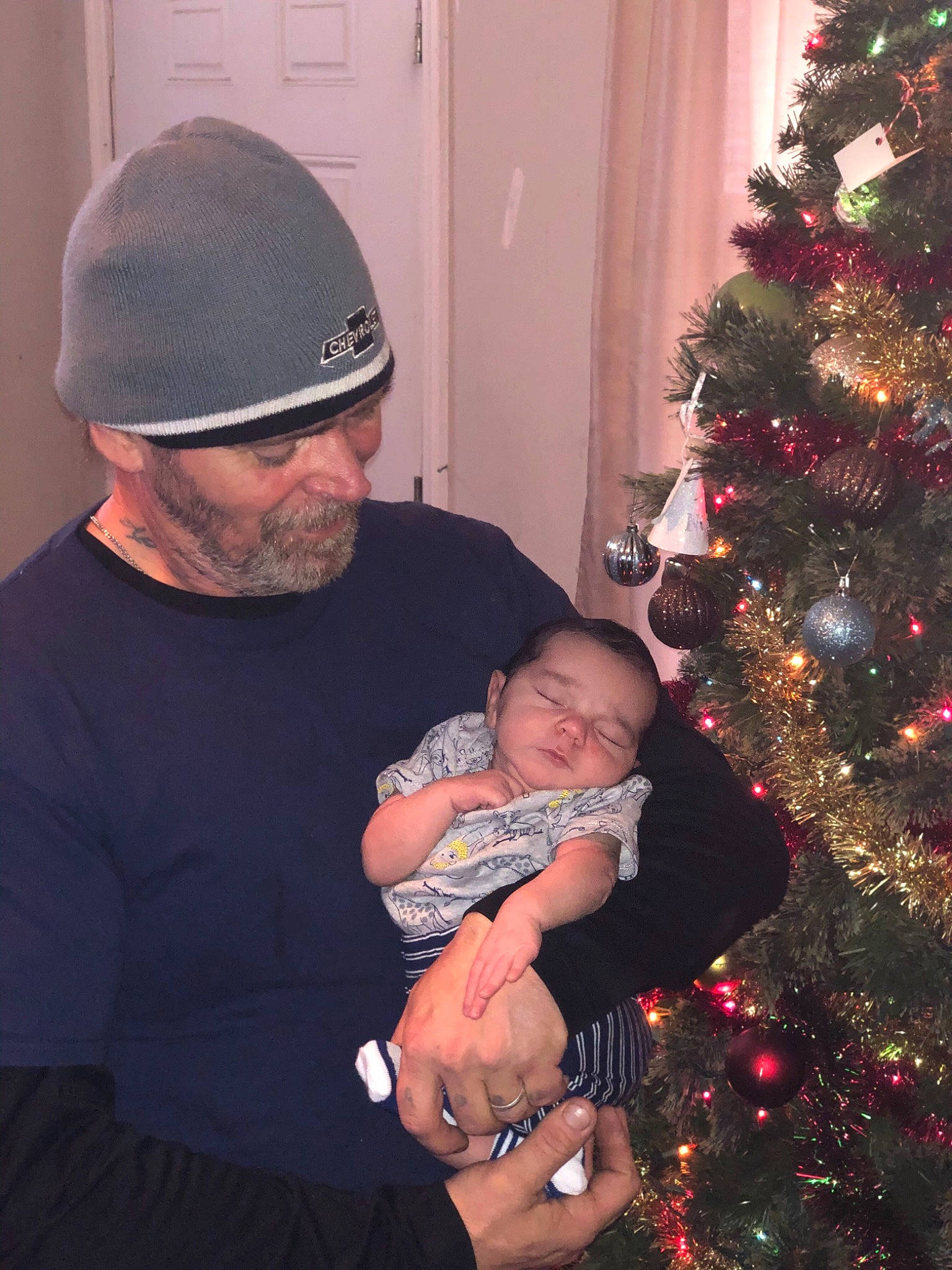 Maverick is registered to the contest to win money with this photo: child, christmas, christmas_decoration, christmas_eve, christmas_ornament, christmas_tree, event, grandparent, holiday, interior_design, lap, person, smile, toddler, tree