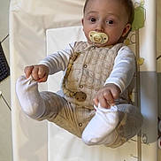 Augustin participe au concours pour gagner de l'argent avec cette photo : baby, infant, pacifier, hands, feet, clothing, changing_pad, floor, tile, cute, child, person, indoor, looking, expression, soft_lighting, white_socks, beige_outfit, holding_feet, playful_design