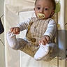 baby, infant, pacifier, hands, feet, clothing, changing_pad, floor, tile, cute, child, person, indoor, looking, expression, soft_lighting, white_socks, beige_outfit, holding_feet, playful_design
