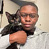 Mittens is registered to the contest to win money with this photo: person, cat, selfie, portrait, indoors, hugging, black_cat, white_paws, whiskers, yellow_eyes, glasses, hoodie, smiling, closeup, pet, companion, human, animal, casual, home