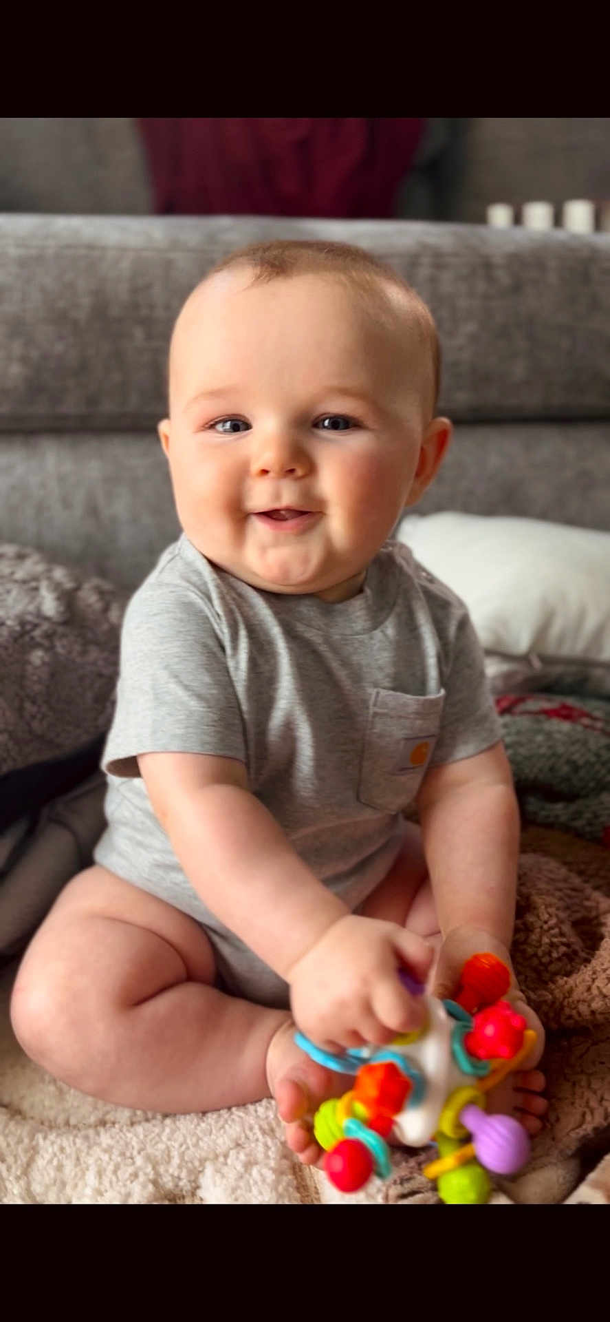 Reece joined the competition — help win amazing prizes! baby, infant, toddler, smiling, sitting, toy, colorful_toy, onesie, gray_shirt, chubby_cheeks, couch, blanket, indoor, portrait, cute, hand, feet, play, face, eyes
