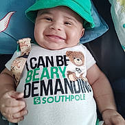 Elijah is registered to the contest to win money with this photo: baby, child, infant, smiling, happy, portrait, green_hat, clothing, teddy_bear, onesie, textile, bed, blanket, indoors, close_up, face, hands, arms, seated, cute