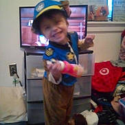 Sawyer Ryan is registered to the contest to win money with this photo: child, costume, police, dog, toy, smile, indoor, television, furniture, clutter, room, floor, sock, plastic_drawer, wall, power_outlet, person, clothing, headwear, play