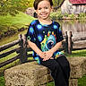 child, smiling, sitting, stone_bench, colorful_shirt, outdoor, fence, pond, barn, grass, tree, nature, portrait, casual_clothing, happy, young_person, scenic, daylight, greenery, relaxed