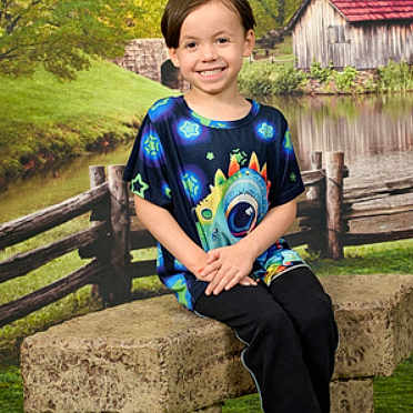 Owen Ernest is registered to the contest to win money with this photo: child, smiling, sitting, stone_bench, colorful_shirt, outdoor, fence, pond, barn, grass, tree, nature, portrait, casual_clothing, happy, young_person, scenic, daylight, greenery, relaxed