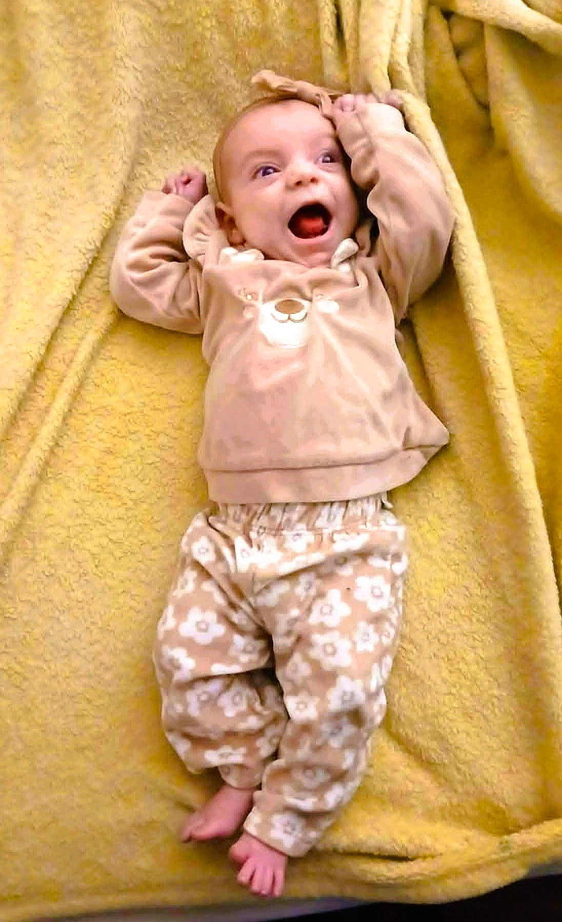 Lilah Jane joined the competition — help win amazing prizes! baby, barefoot, blanket, child, clothing, cute, floral_pattern, happy, indoors, infant, lying_down, pants, playful, portrait, raised_arms, small_feet, smiling, soft_texture, sweatshirt, yellow