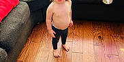 Koa is registered to the contest to win money with this photo: toddler, child, barefoot, shirtless, pants, wooden_floor, couch, living_room, cushion, clothing, home_interior, natural_light, curly_hair, standing, expression, person, floor, furniture, casual, indoors