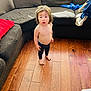toddler, child, barefoot, shirtless, pants, wooden_floor, couch, living_room, cushion, clothing, home_interior, natural_light, curly_hair, standing, expression, person, floor, furniture, casual, indoors