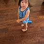 child, barefoot, blue_tank_top, standing, wooden_floor, indoors, curious_expression, toddler, hair, person, floor, toy, casual_clothing, looking_up, young_child, home, skin, face, solemn, standing_barefoot