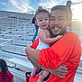 baby, man, child, bleachers, outdoor, sky, clouds, smile, happy, tattoo, red_shirt, orange_shirt, sunglasses, high_school, people, daylight, fence, family, holding, portrait