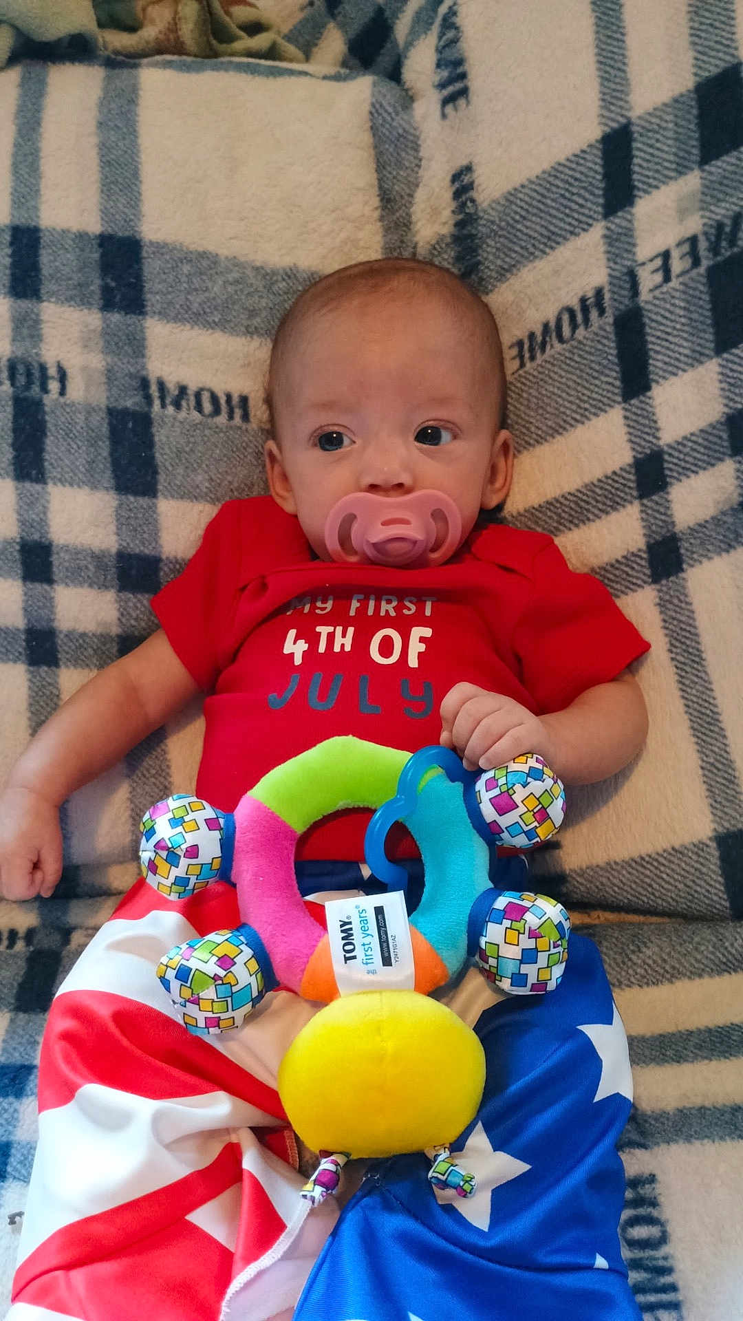 Penelope is registered to the contest to win money with this photo: baby, infant, pacifier, plush_toy, red_clothing, blue_clothing, american_flag_pattern, blanket, lying_down, cute, child, celebration, holiday_outfit, colorful, patterned, indoor, person, portrait, small_hand, cozy