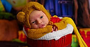 élie a rejoint le concours — aidez-le/la à gagner de superbes lots ! baby, infant, knit_hat, yellow_clothing, red_bucket, white_cushion, face, portrait, cute, resting, indoors, colorful_background, child, newborn, soft_texture, warm_clothing, headgear, expression, small_hands, cozy