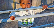 David Afanbo a rejoint le concours — aidez-le/la à gagner de superbes lots ! toddler, child, metal_fence, blue, rust, concrete_wall, outdoor, sunlight, shadow, curious, grip, casual_clothing, long_sleeve_shirt, daylight, urban, expression, person, young_child, standing, playful
