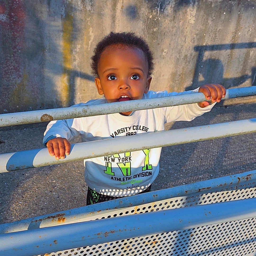 David Afanbo a rejoint le concours — aidez-le/la à gagner de superbes lots ! toddler, child, metal_fence, blue, rust, concrete_wall, outdoor, sunlight, shadow, curious, grip, casual_clothing, long_sleeve_shirt, daylight, urban, expression, person, young_child, standing, playful