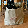 black_cat, cat, paper_bag, bag, cracker_barrel_logo, green_eyes, wooden_floor, hardwood_floor, chair, dining_table, indoor, pet, cute, curious, sitting, portrait, domestic_animal, fur, handle, household_object
