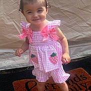 Ellie is registered to the contest to win money with this photo: toddler, child, pink_bow, pink_outfit, strawberry_design, sandals, welcome_mat, smiling, standing, outdoor, grass, sunlight, happy, cute, portrait, person, clothing, footwear, hair, background