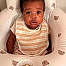 baby, infant, child, portrait, sitting, wide_eyes, curly_hair, facial_expression, bib, striped_clothing, pink_clothing, baby_seat, nursery, cushion, bear_pattern, indoor, warm_lighting, closeup, adorable, photography