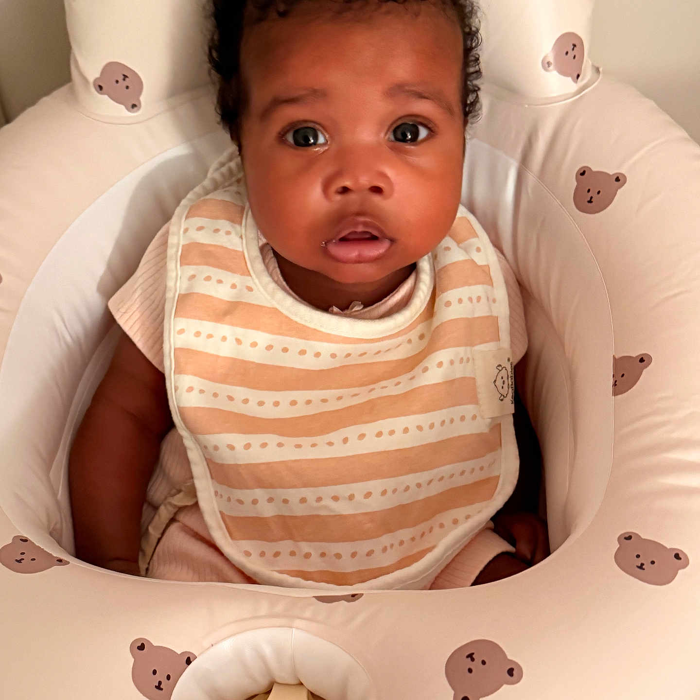Alayna joined the competition — help win amazing prizes! baby, infant, child, portrait, sitting, wide_eyes, curly_hair, facial_expression, bib, striped_clothing, pink_clothing, baby_seat, nursery, cushion, bear_pattern, indoor, warm_lighting, closeup, adorable, photography