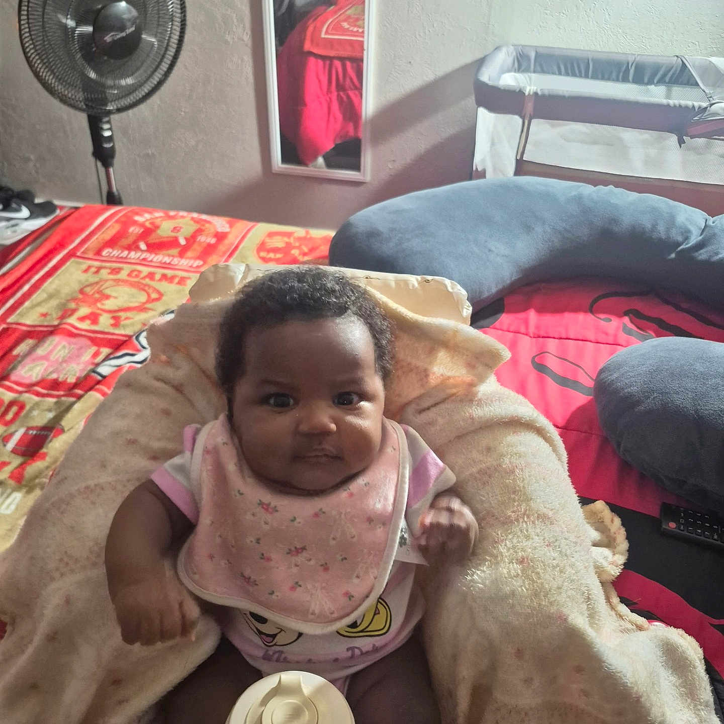 Isis is registered to the contest to win money with this photo: baby, blanket, bib, bottle, bed, fan, mirror, crib, pillow, clothing, indoor, person, infant, room, soft_toy, furniture, child, cozy, portrait, seated