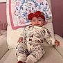 baby, smile, pajamas, headband, red_bow, armchair, blanket, floral_pattern, pink_wall, cozy, indoors, child, cute, socks, holiday_theme, furniture, person, infant, happy, portrait