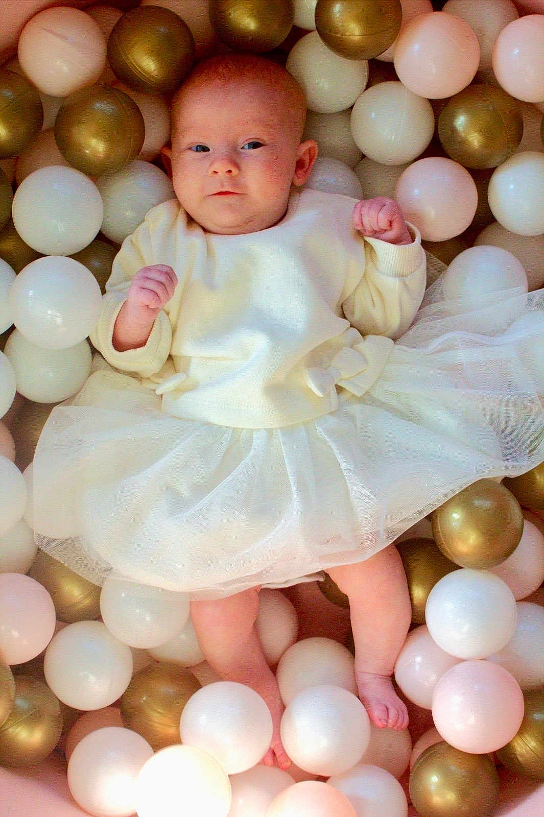 Pennie-Mae joined the competition — help win amazing prizes! baby, infant, white_dress, ball_pit, plastic_balls, gold_balls, pastel_colors, cute, portrait, child, soft_lighting, indoor, playful, fists, newborn, skin, feet, eyes, head, clothing