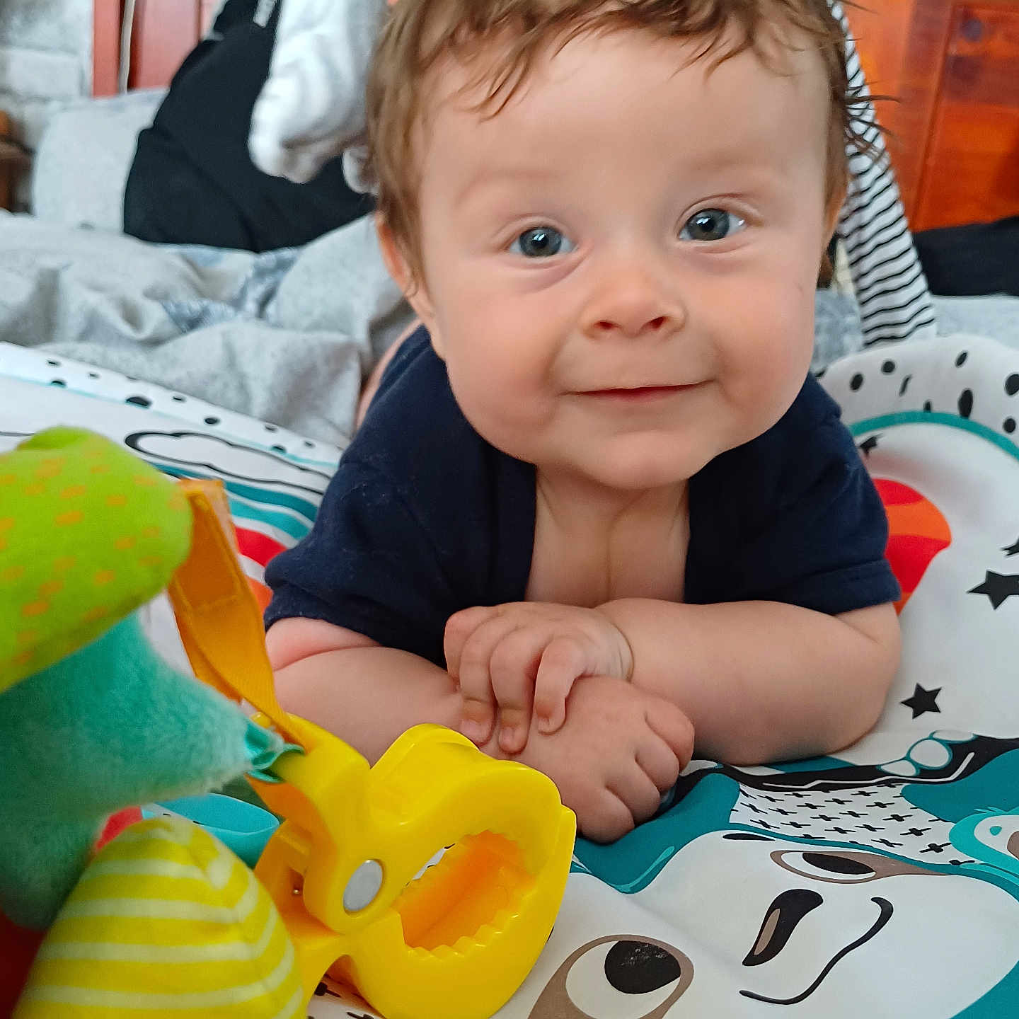 Oliver is registered to the contest to win money with this photo: baby, blanket, blue_eyes, child, closeup, colorful, cute, face, hands, happy, indoor, infant, patterned_mat, play_mat, smiling, soft_toys, striped_clothing, toy, wooden_headboard, yellow_toy