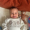 baby, smiling, infant, teddy_bear, toy, blanket, sweater, penguin_design, mat, plaything, child, cute, indoors, happy, portrait, lying_down, clothing, face, hand, warm_colors