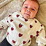 amour, baby, black_pants, casual_clothing, child, comfortable, cute, face, happy, indoors, infant, lying_down, portrait, red_hearts, smiling, soft_texture, towel, warm_colors, white_sweater, young_child