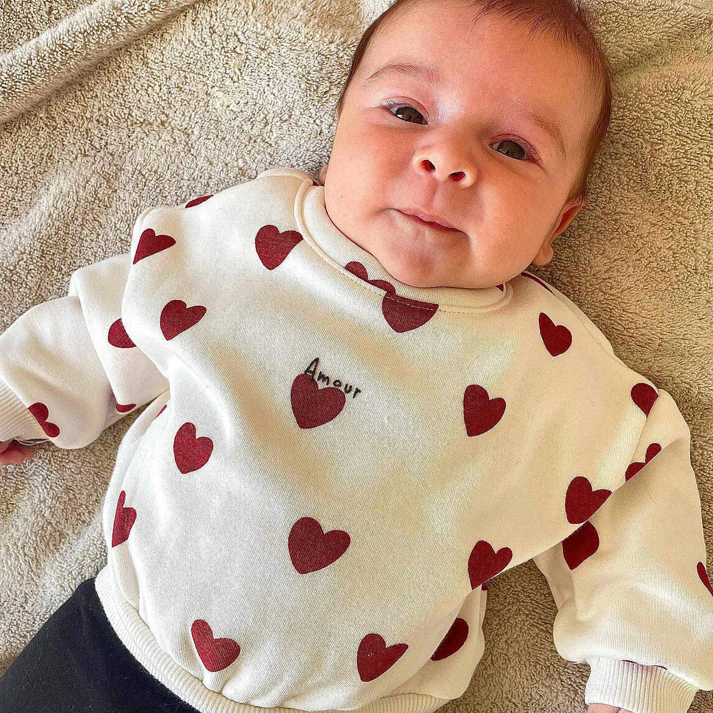 Ophélya a rejoint le concours — aidez-le/la à gagner de superbes lots ! amour, baby, black_pants, casual_clothing, child, comfortable, cute, face, happy, indoors, infant, lying_down, portrait, red_hearts, smiling, soft_texture, towel, warm_colors, white_sweater, young_child