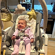 Andie is registered to the contest to win money with this photo: baby, infant, smiling, baby_rocker, baby_toys, mobile, plush_toys, moon, star, cloud, pink_clothing, kitchen, appliances, toaster, microwave, stove, wooden_floor, cushion, harness, happy