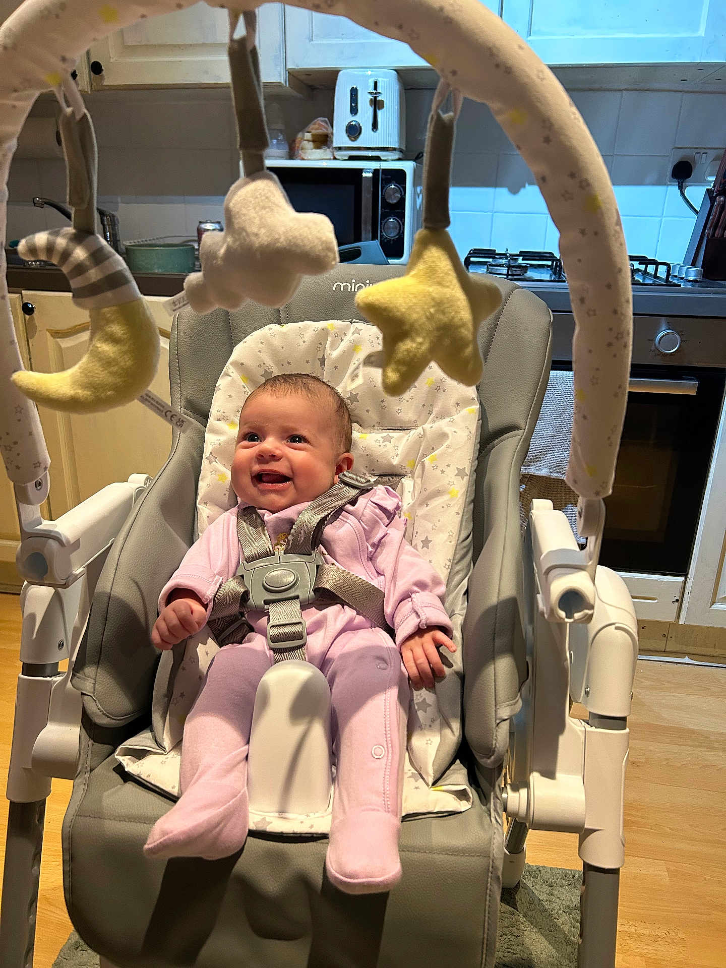 Andie is registered to the contest to win money with this photo: baby, infant, smiling, baby_rocker, baby_toys, mobile, plush_toys, moon, star, cloud, pink_clothing, kitchen, appliances, toaster, microwave, stove, wooden_floor, cushion, harness, happy