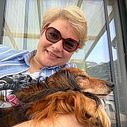 Guess participe au concours pour gagner de l'argent avec cette photo : person, dog, smile, sunglasses, outdoor, window, glass_door, pet, cuddle, happy, long_hair, collar, casual_clothing, relaxed, portrait, daylight, home, companion, friendly, close_up