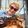 person, dog, smile, sunglasses, outdoor, window, glass_door, pet, cuddle, happy, long_hair, collar, casual_clothing, relaxed, portrait, daylight, home, companion, friendly, close_up