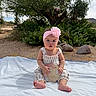 baby, infant, child, outdoor, blanket, flower_headband, sign, nature, greenery, rocks, tree, desert, sunlight, sitting, cute, portrait, clothing, pink, white, person