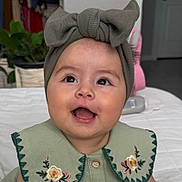 Layla joined the competition — help win amazing prizes! baby, child, smiling, headband, bow, collar, embroidered, green_clothing, portrait, indoor, table, plant, face, happy, cute, person, apparel, infant, closeup, expression