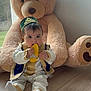 baby, chewing, child, cute, daylight, floor, hat, home, indoor, large_toy, person, plush, sitting, stuffed_animal, teddy_bear, toy, traditional_clothing, window, wooden_floor, yellow_shoe
