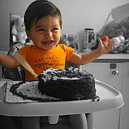 Anthony joined the competition — help win amazing prizes! toddler, child, high_chair, cake, smiling, happy, orange_shirt, food, celebration, birthday, indoors, table, face, arms_raised, feet, photography, monochrome_background, baby, sitting, fun
