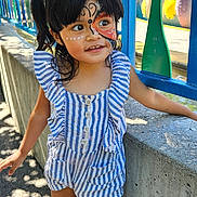 Maryann is registered to the contest to win money with this photo: child, face_paint, outdoor, daylight, striped_clothing, romper, sneakers, concrete_wall, colorful_fence, blue, green, curly_hair, smiling, playground, sunlight, shadow, person, young_child, happy, casual_wear