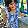 child, face_paint, outdoor, daylight, striped_clothing, romper, sneakers, concrete_wall, colorful_fence, blue, green, curly_hair, smiling, playground, sunlight, shadow, person, young_child, happy, casual_wear