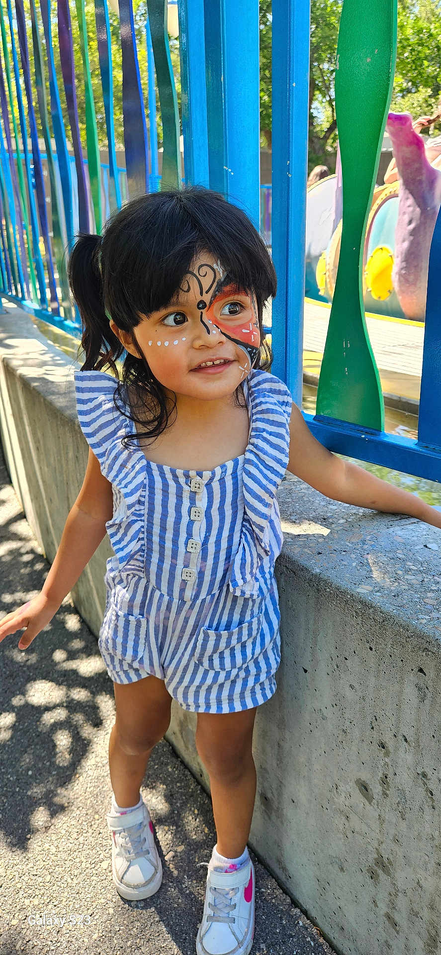 Maryann is registered to the contest to win money with this photo: child, face_paint, outdoor, daylight, striped_clothing, romper, sneakers, concrete_wall, colorful_fence, blue, green, curly_hair, smiling, playground, sunlight, shadow, person, young_child, happy, casual_wear
