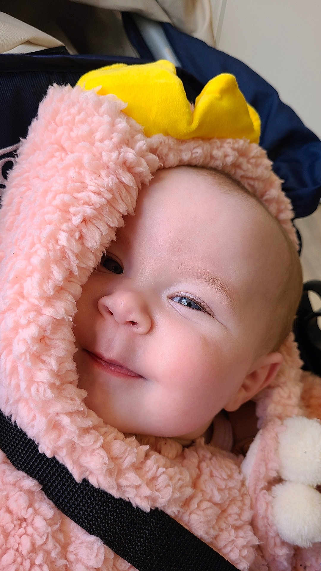 Nina participe au concours pour gagner de l'argent avec cette photo : baby, infant, smile, close_up, pink_fur, hood, pom_poms, navy_background, yellow_hat, cheeks, eyes, nose, lips, soft_texture, stroller, safety_strap, portrait, cozy, clothing, adorable