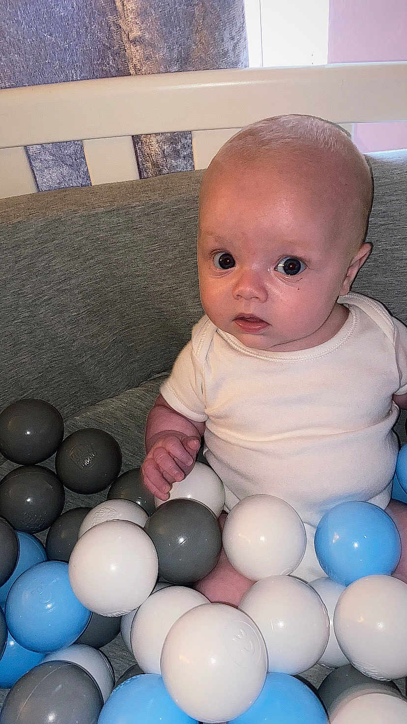Charlie is registered to the contest to win money with this photo: baby, child, infant, ball_pit, plastic_balls, crib, white_clothing, blue_balls, gray_balls, indoor, person, cute, seated, looking, curious, face, head, small_hand, soft_lighting, closeup