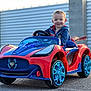 toddler, child, toy_car, red, blue, smiling, outdoor, pavement, superhero, hoodie, vehicle, wheels, daylight, happy, small_child, person, play, fun, young, casual