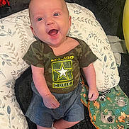 Henry is registered to the contest to win money with this photo: baby, smiling, happy, infant, camo_shirt, pillow, blanket, animal_print, child, indoors, cute, sitting, clothing, face, person, young_child, expression, joy, comfort, fabric